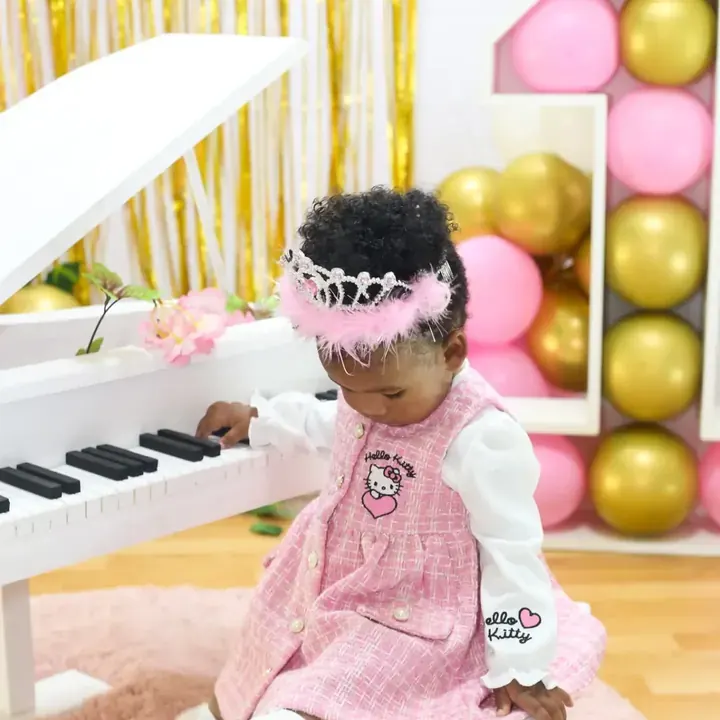 First birthday session, a baby girl in a pink pinafore and tiara at a white toy piano