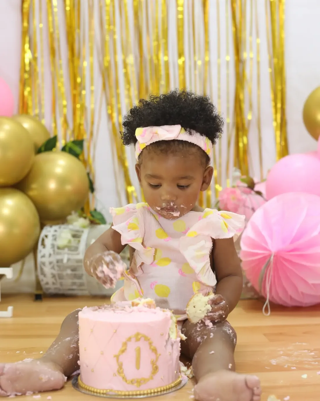First birthday cake-smash session, a baby girl digging into a pink number-one cake on a gold and pink setup